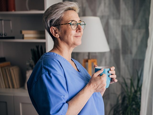 Person in medizinischer Arbeitskleidung steht in einem Innenraum und hält eine Tasse, als Symbol für Ruhe, Stabilität und finanzielle Planung im Berufsalltag von Heilberuflern.