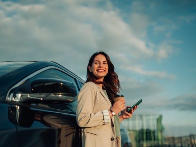 Frau mit Handy und Coffee-to-go lehnt an einer schwarzen Limousine
