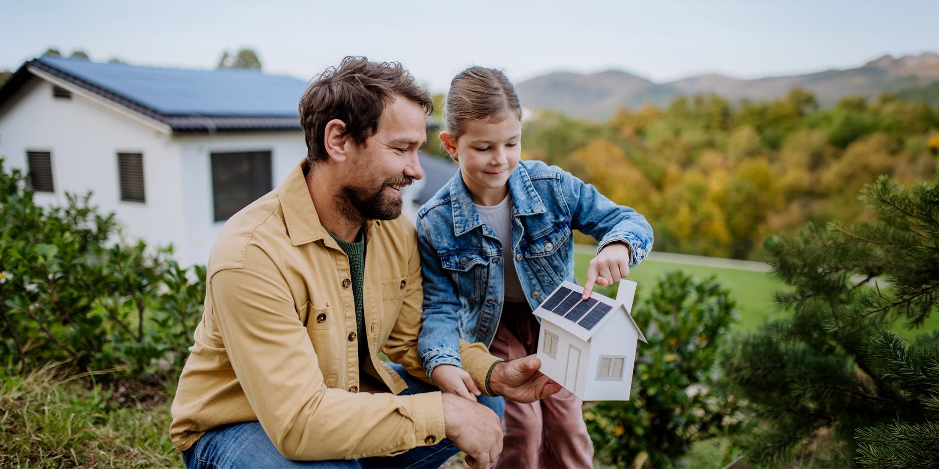 Vater und Tochter blicken lächelnd auf das Modell eines Einfamilienhauses mit Photovoltaikmodulen auf dem Dach.