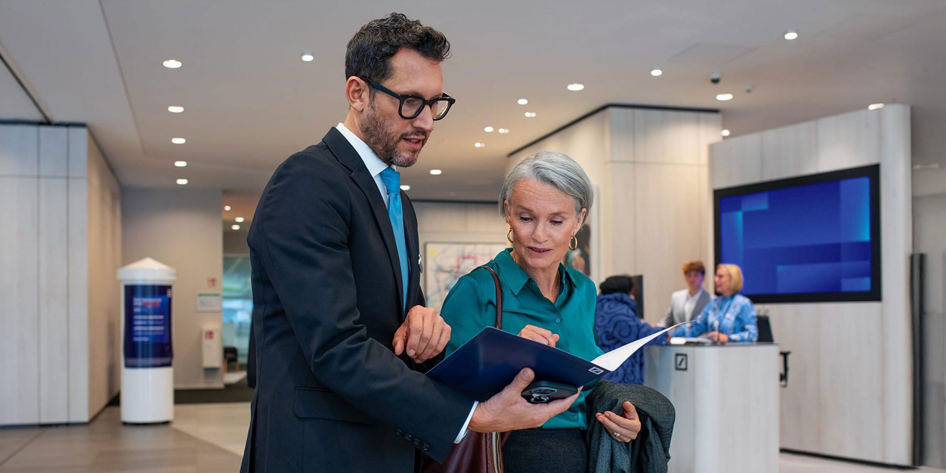 Berater der Deutschen Bank mit Unterlagenmappe in der Hand im Gespräch mit einer Kundin in der Filiale.