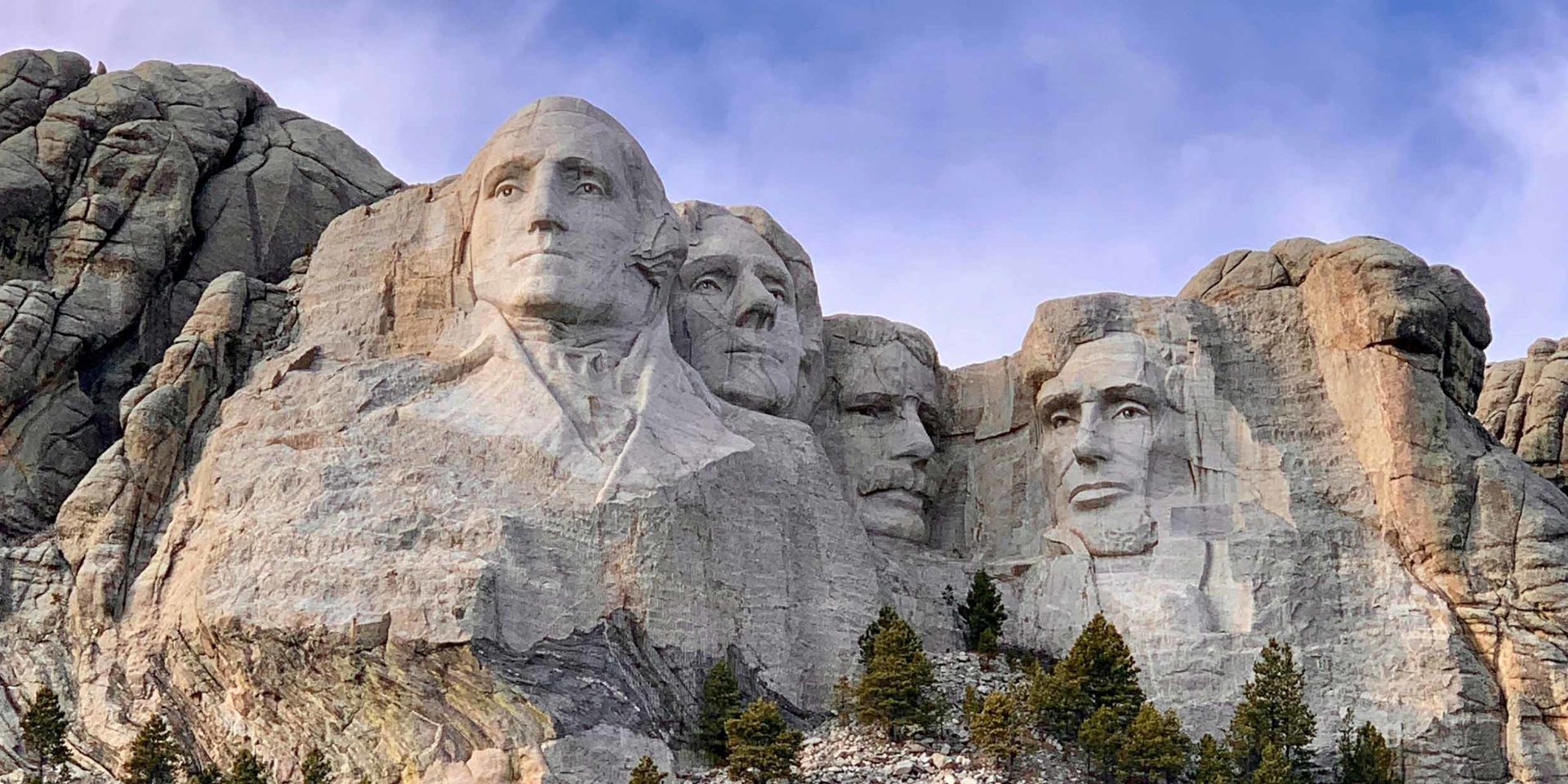 Mount Rushmore in South Dakota mit den in den Granit gemeißelten Porträts der US-Präsidenten George Washington, Thomas Jefferson, Theodore Roosevelt und Abraham Lincoln vor blauem Himmel.