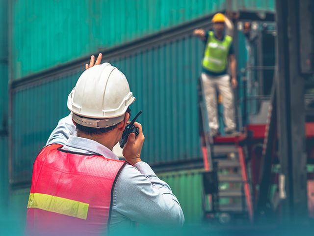 Ein Mann mit Warnweste und Helm dirigiert einen Gabelstaplerfahrer per Walkytalky